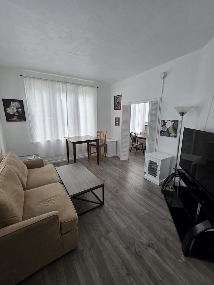 Living room with a beige sofa, coffee table and television, a dining table beside a curtained window, and wood-look plank flooring