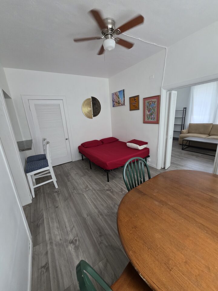 Room with a ceiling fan, a red day bed, a round wooden dining table with green chairs and a louvered closet, opening through to the living room