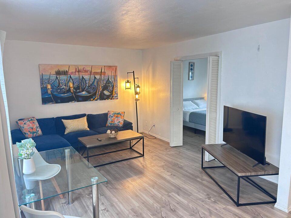Living room with a navy sectional sofa, framed artwork and a glass table, bedroom beyond