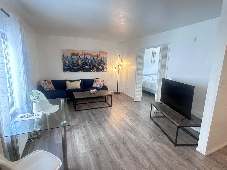 Living room with a navy sectional beneath a painting of Venetian gondolas, a wood and metal coffee table, a standing lamp, a television on a matching console and a glass dining table by the window, on grey wood-look floors, with louvered doors open to the bedroom