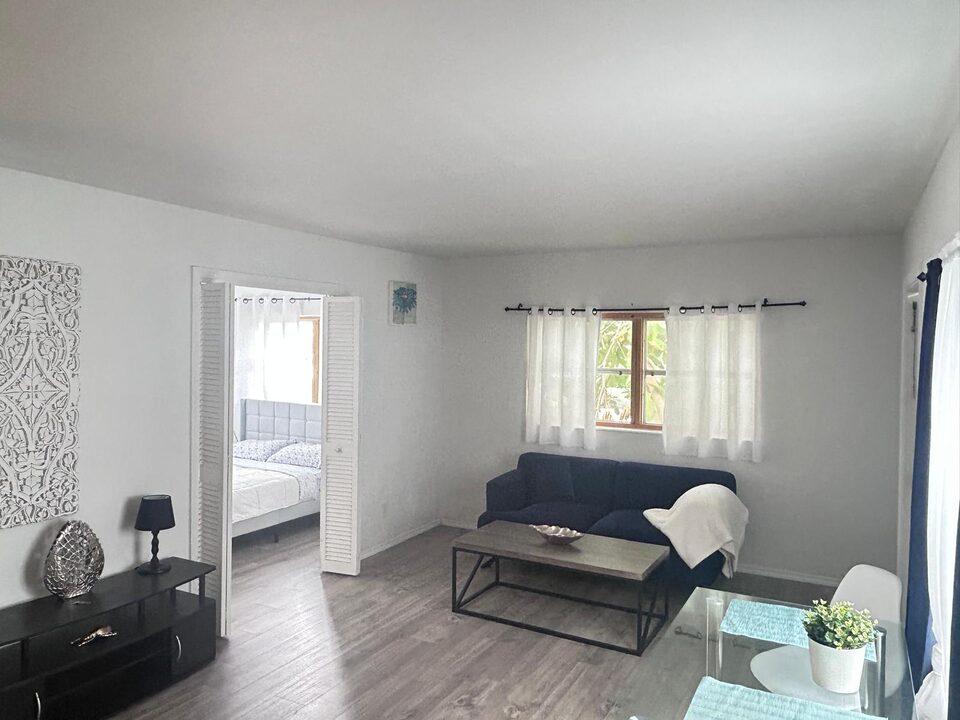 Living room with a navy sofa and a white throw beneath a curtained window, a wood-top coffee table, a black media console with a lamp and a carved white wall panel, looking through open louvered doors to the bedroom, with a glass dining table in the foreground, on grey wood-look plank flooring