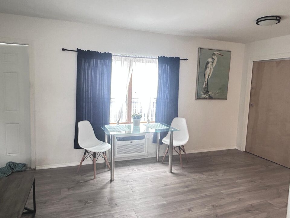 Dining area with a glass table and two white moulded chairs beneath a window with navy curtains, a wall air conditioner under the window, a heron painting and the front door