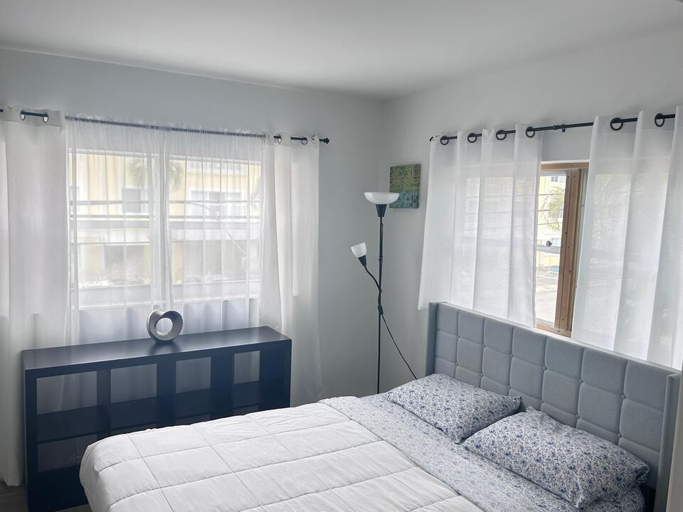 Bedroom with a grey upholstered headboard, a made-up bed with blue floral pillows, sheer curtains at two windows, a black cube shelf and a floor lamp