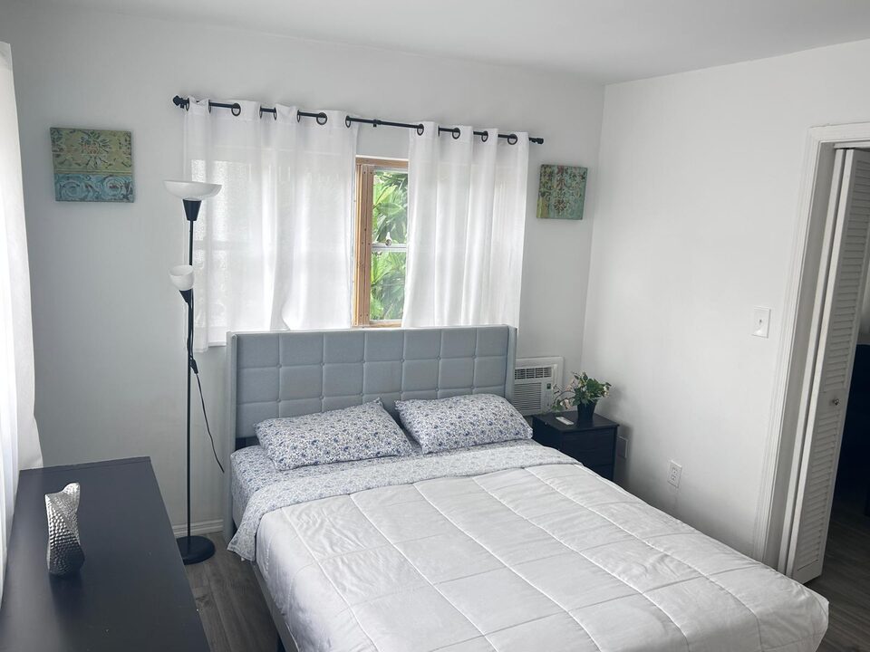 Bedroom seen from the other side of the bed, with the grey upholstered headboard beneath a curtained window, a wall air conditioner, a nightstand with a plant, a black dresser and louvered doors to the living room