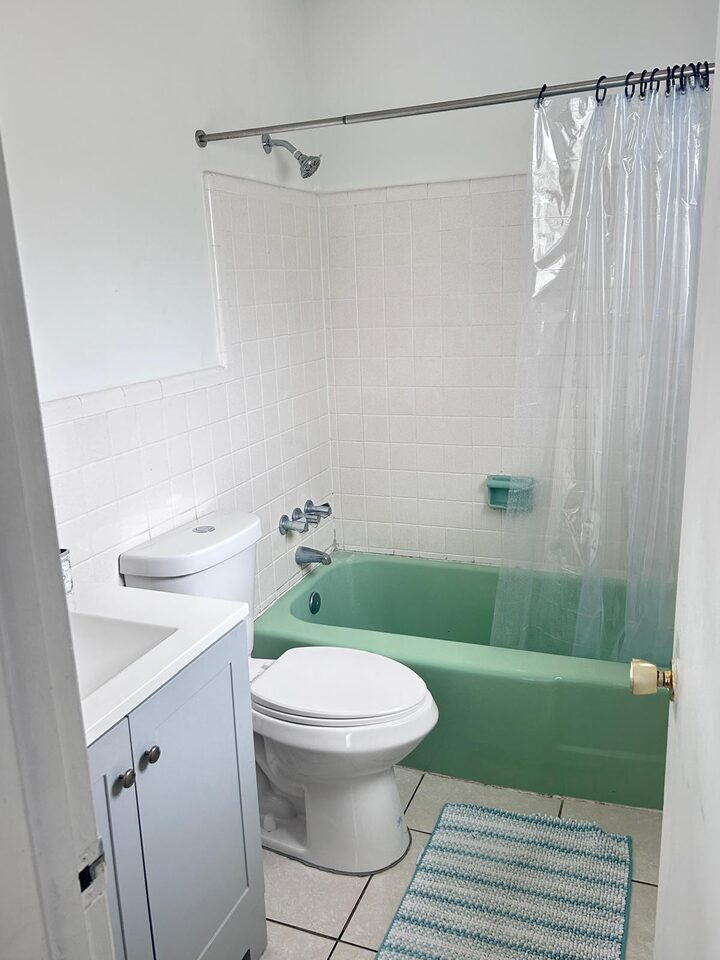 Bathroom with a green tub behind a clear shower curtain, white tiled walls, a toilet, a grey vanity and a striped bath mat on white tile