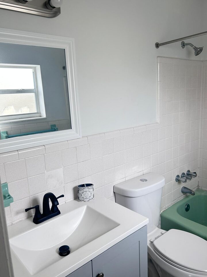 Bathroom vanity with a white basin and a black tap, a white-framed mirror and white tiled walls, with the toilet and the green tub beyond