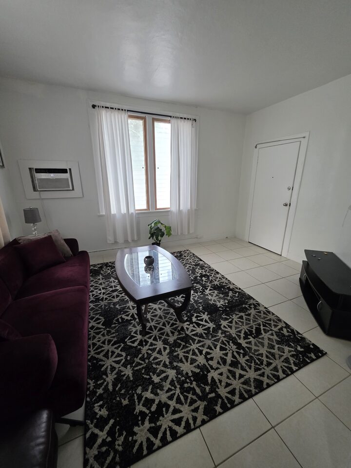 Living room with a burgundy velvet sofa and a glass coffee table on a patterned rug, with white tile floors and a curtained window