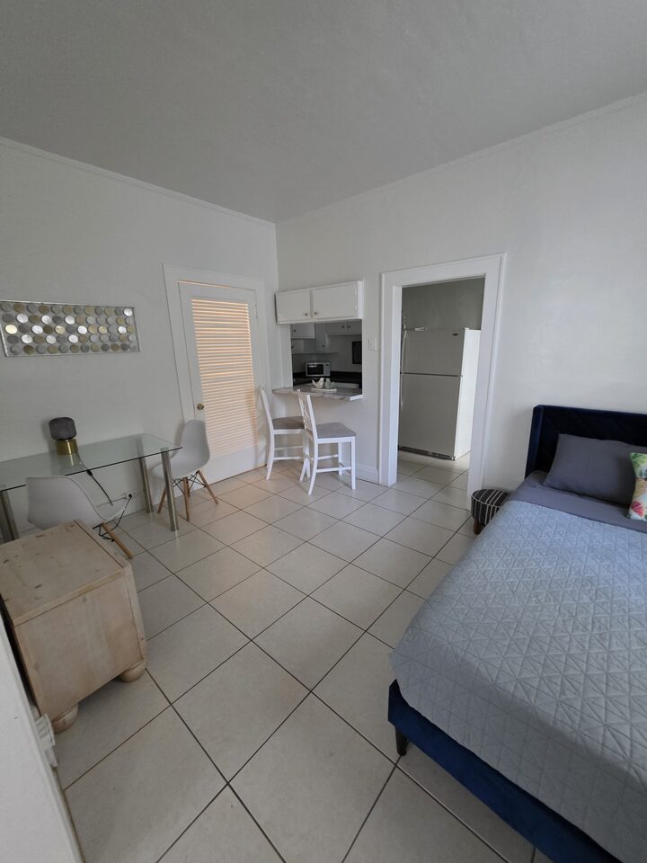 Bedroom with a navy upholstered bed and a glass dining table, with the kitchen visible through the doorway