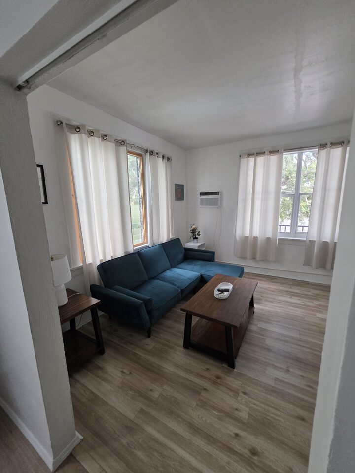 Living room with a teal velvet sectional sofa, a wooden coffee table, two curtained windows and wood-look plank flooring