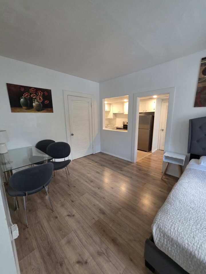 Bedroom with an upholstered headboard and a glass dining table, with the kitchen beyond the pass-through