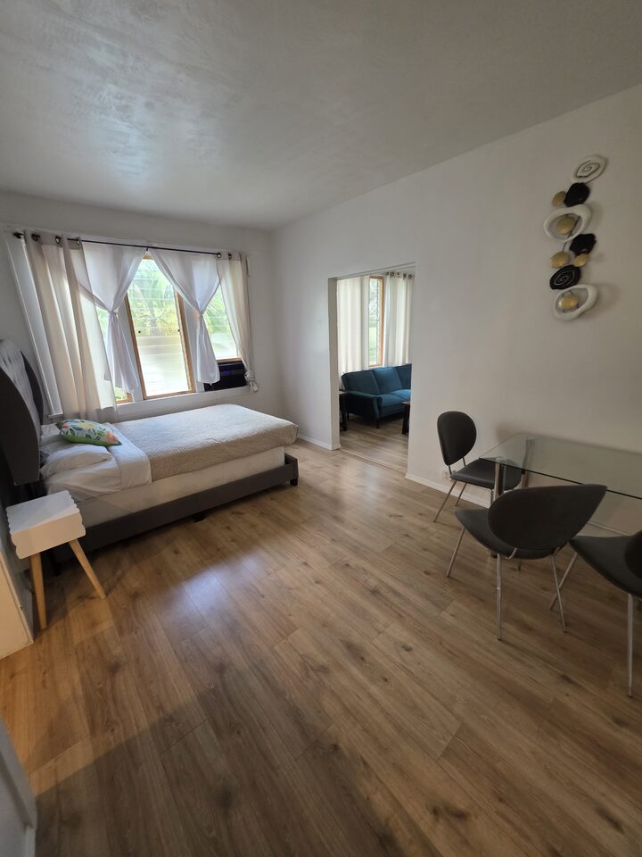 Bedroom with a curtained window and a bedside table, looking through to the living room