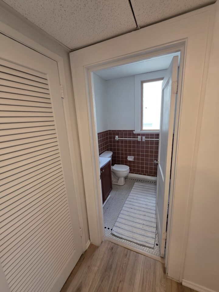 Hallway with a louvered closet door, looking into the bathroom with its tiled wainscot