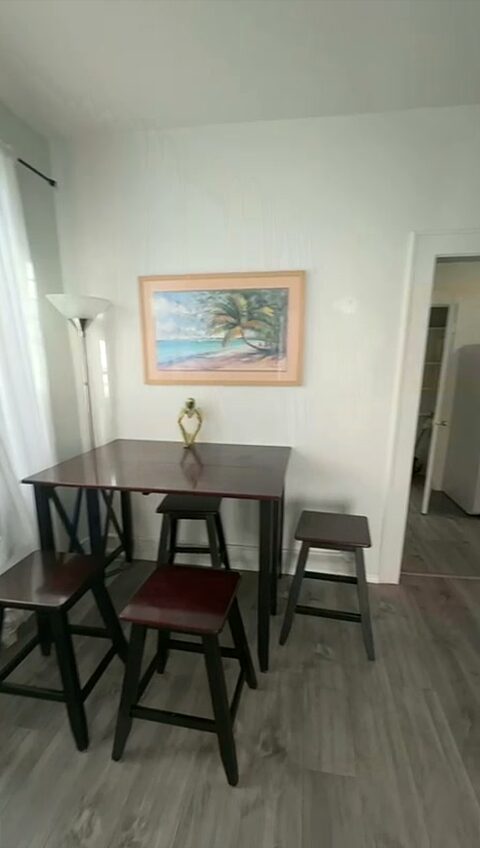 Dining area with a dark wood high table, three stools, a framed beach print and a sheer-curtained window