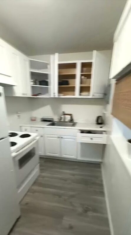 Galley kitchen with a white electric range, full-height refrigerator, stainless steel sink, white cabinets and glass-fronted upper cupboards