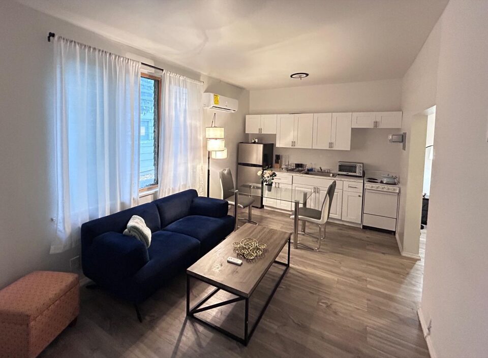 Living room with a navy sofa beneath a sheer-curtained window, an orange patterned ottoman, a wood and metal coffee table and a glass dining table with two grey chairs, opening onto a kitchen of white cabinets with a stainless steel refrigerator and an electric range, on grey wood-look floors