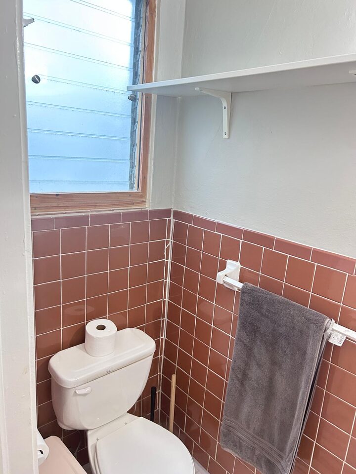Bathroom with a toilet, terracotta tiled walls, a shelf above and a towel rail, beside an obscured-glass louvered window