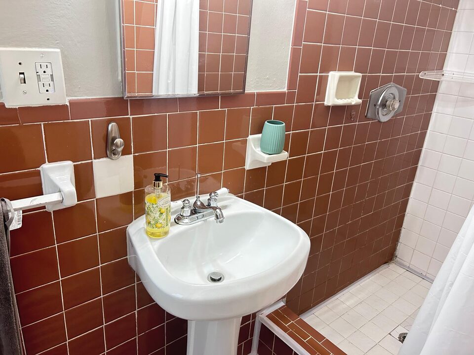 Bathroom with a pedestal sink, a chrome-framed mirror and terracotta tiled walls, with the shower beyond