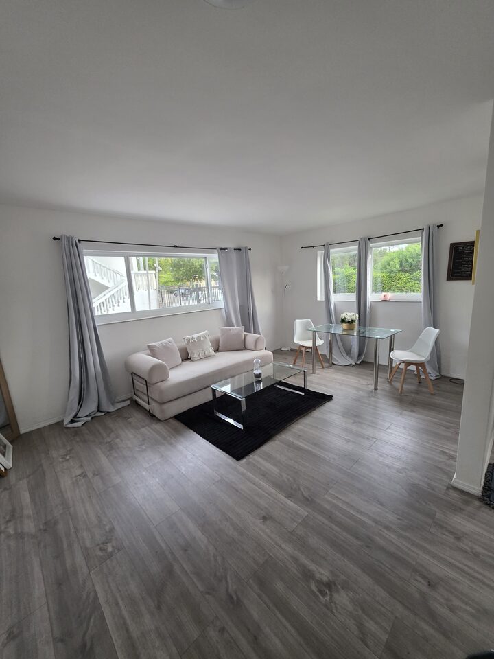 Living room with a cream sofa, a glass coffee table, a glass dining table with white chairs and two curtained windows