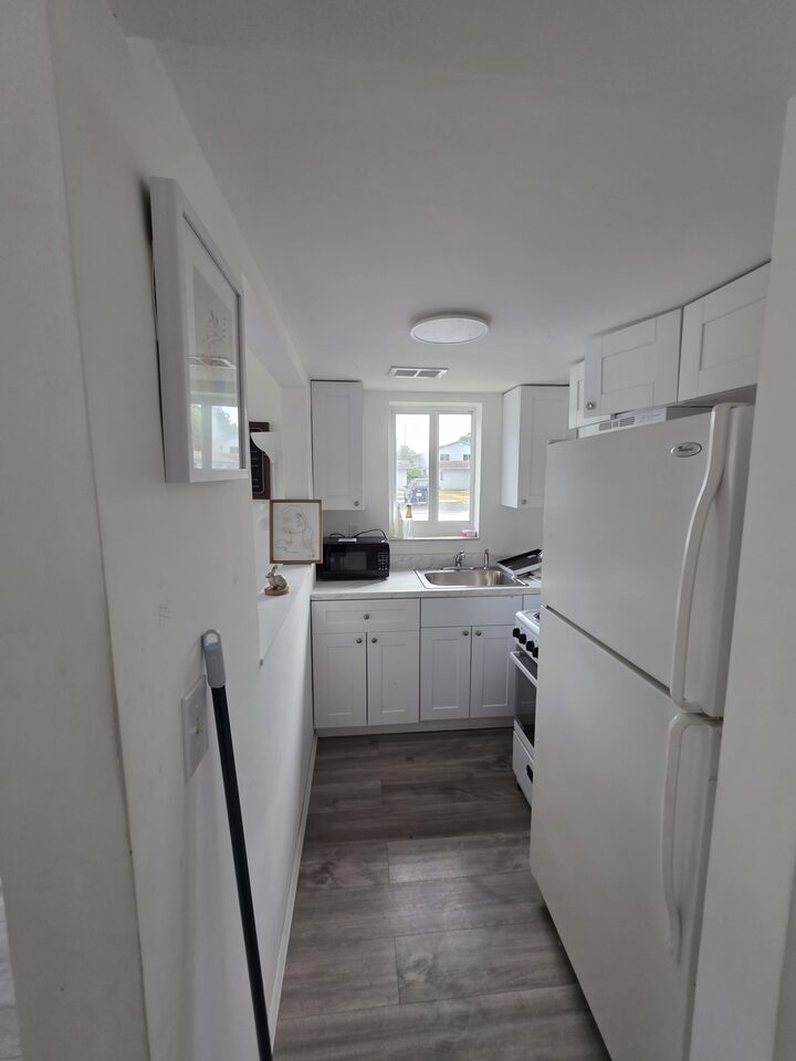 Galley kitchen seen from the hall, with white cabinets, a refrigerator, a microwave and a sink under the window
