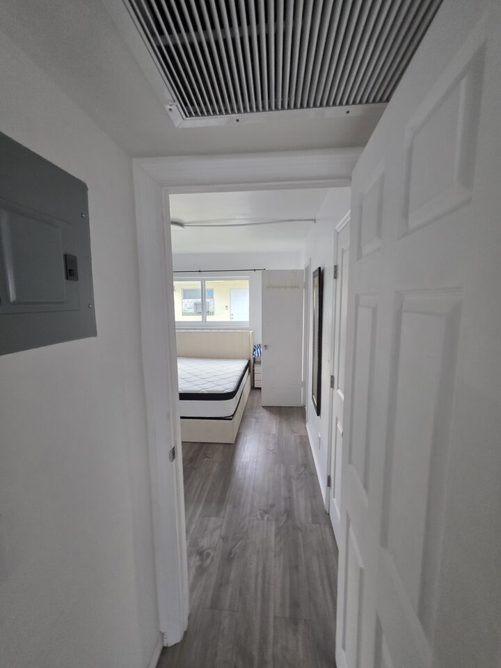 Hallway looking into the bedroom, with plank flooring and a ceiling air vent