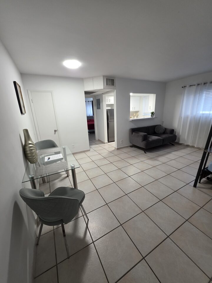 Living and dining area with a glass dining table, a grey loveseat and the kitchen pass-through, on white tile floors