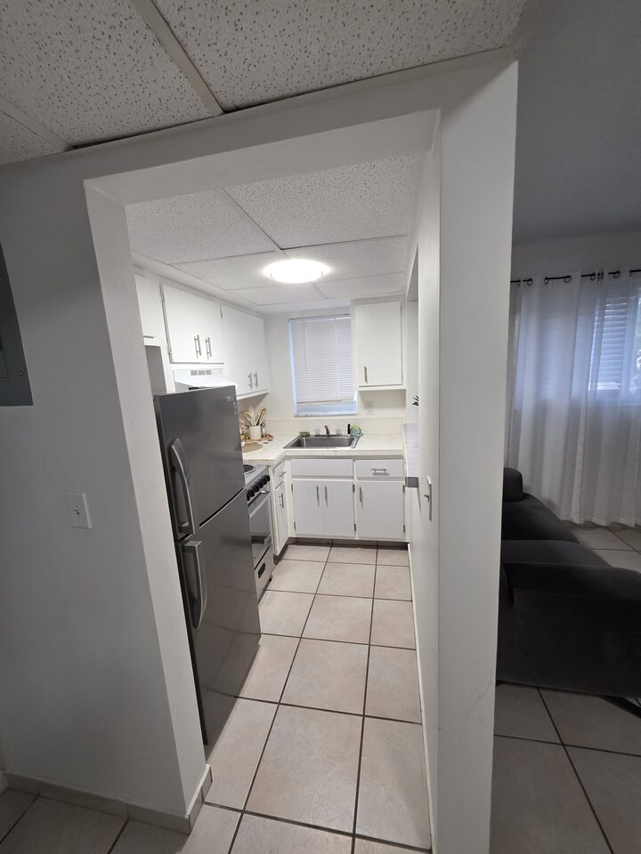 Kitchen seen through the doorway, with white cabinets, a stainless steel refrigerator, an electric range and a sink under the window