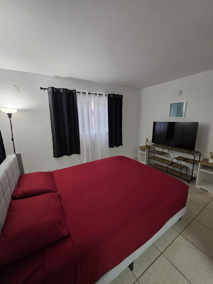 Bedroom with an upholstered headboard, red bedding, blackout curtains and a television on a console