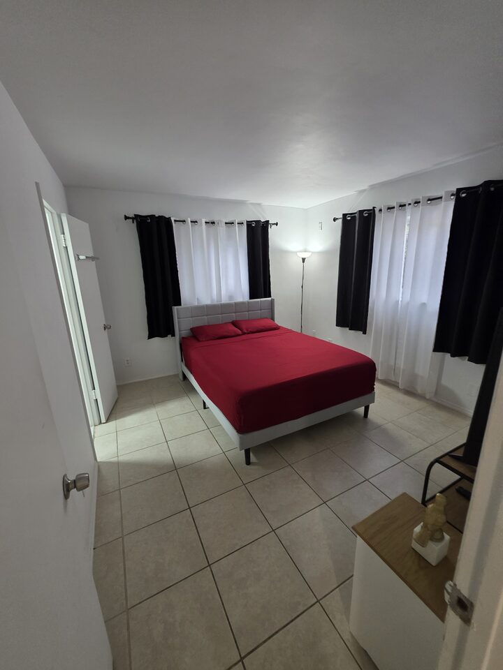 Bedroom seen from the doorway, with the bed, blackout and sheer curtains and a bedside table