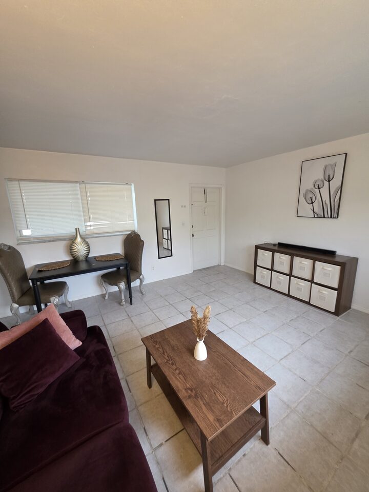 Living room with a dining table and two upholstered chairs by the window, a storage unit with baskets and the front door