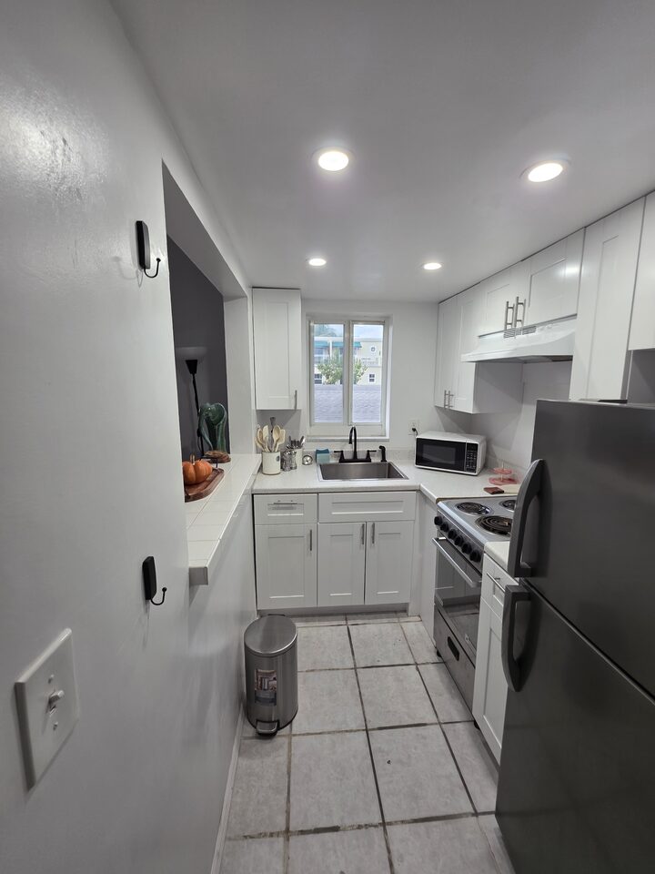 Galley kitchen with white cabinets, a tiled countertop, a stainless steel sink under the window and a pass-through to the living room