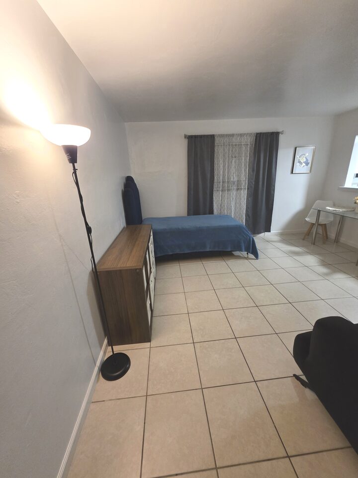 Living area with a bed against the curtained window, a wooden storage unit with fabric bins, a standing lamp and a dining table under the window
