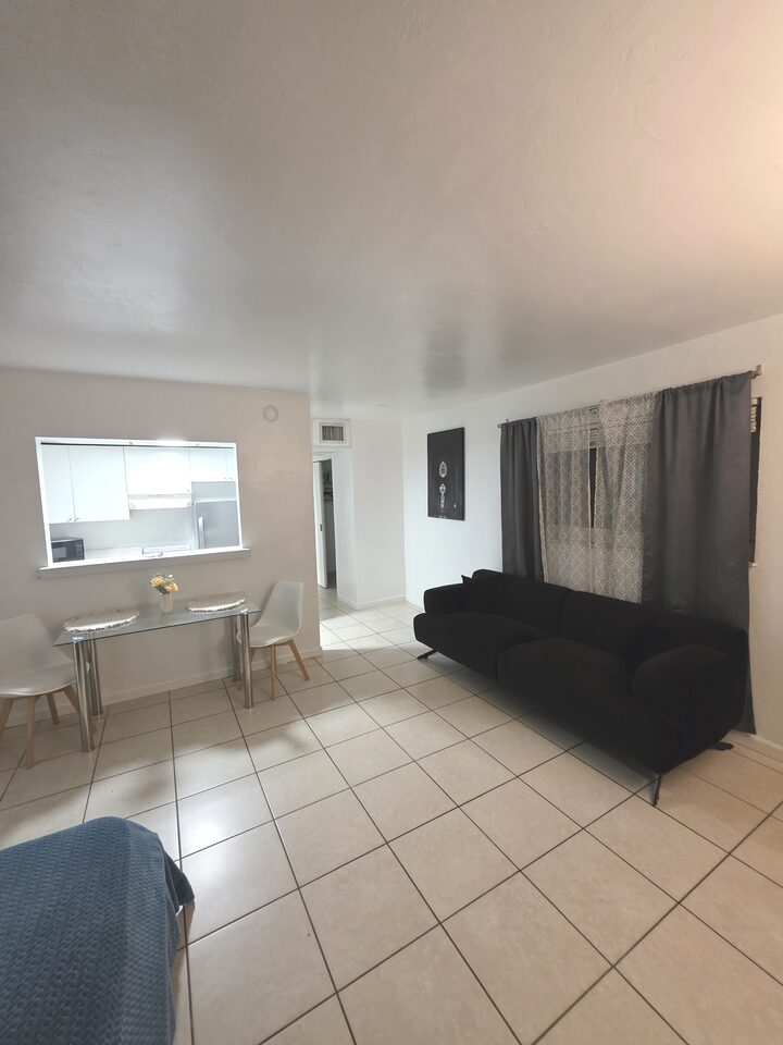 Living and dining area with a black sofa beneath a curtained window, a glass dining table with upholstered chairs, framed art and the kitchen pass-through, on cream tile floors