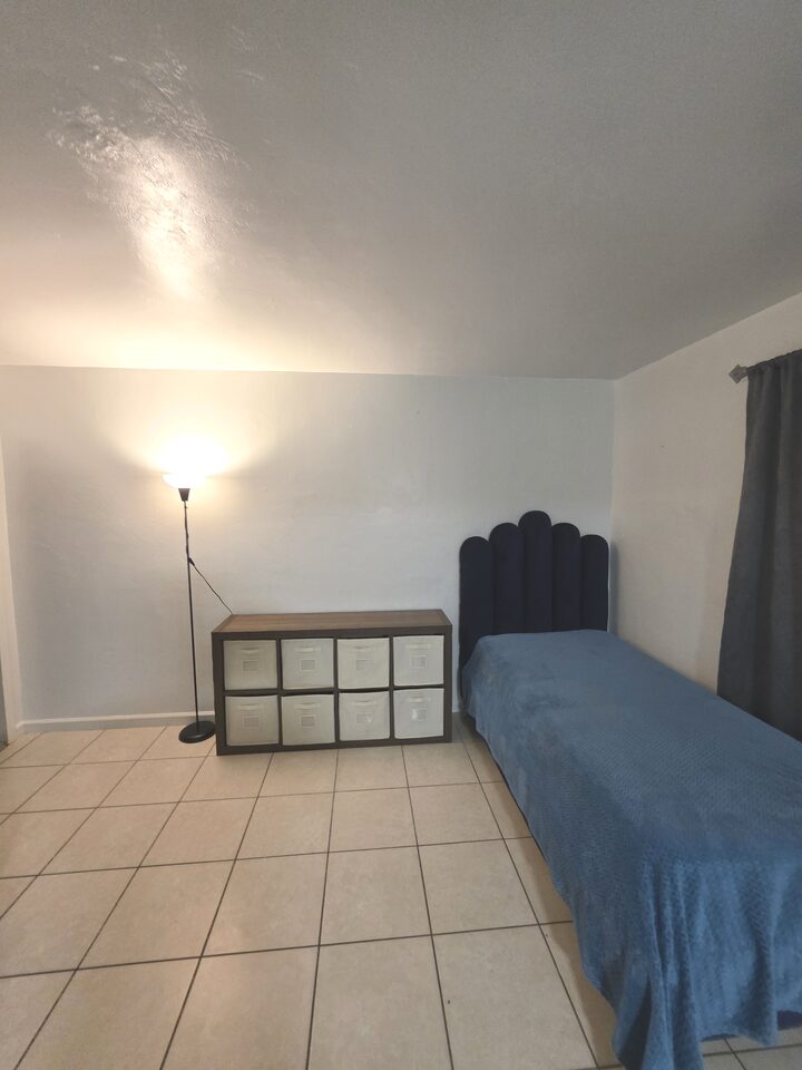 Living area with a bed with a navy channel-tufted headboard, a wooden storage unit with fabric bins and a standing lamp, on cream tile floors