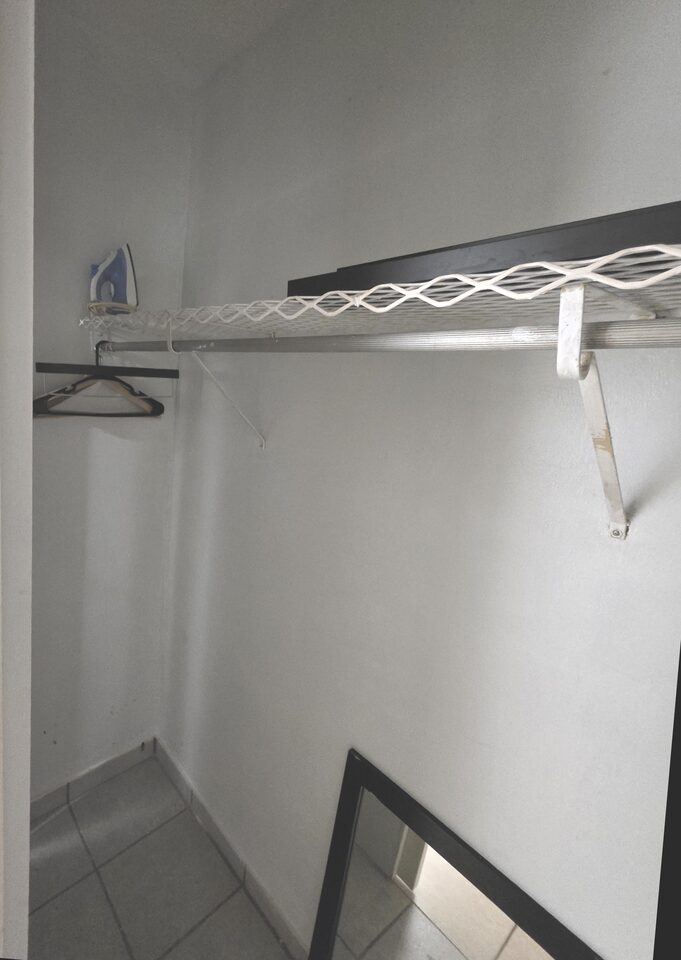 Closet with a wire shelf and hanging rail, an iron on the shelf and a mirror propped against the wall