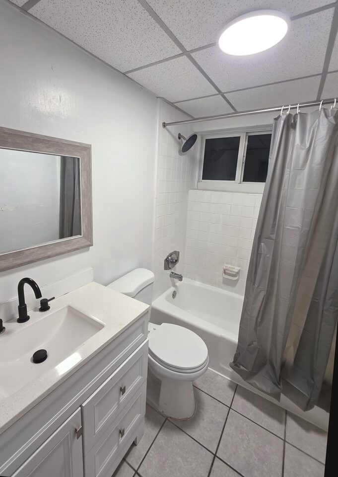 Bathroom with a white vanity and a black tap, a framed mirror, a toilet and a tub behind a shower curtain, with a window above