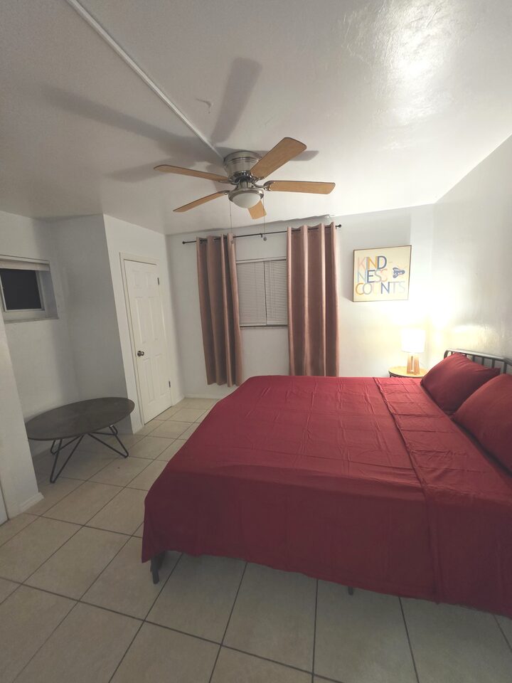 Bedroom with a ceiling fan, a bed in burgundy bedding, curtains drawn back from a blinded window and a lamp on a round bedside table