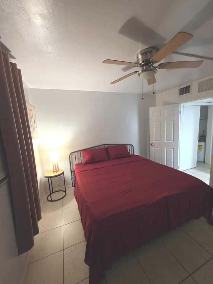 Bedroom seen from the opposite corner, with a black metal bed frame, burgundy bedding, a bedside lamp and the door open to the hallway