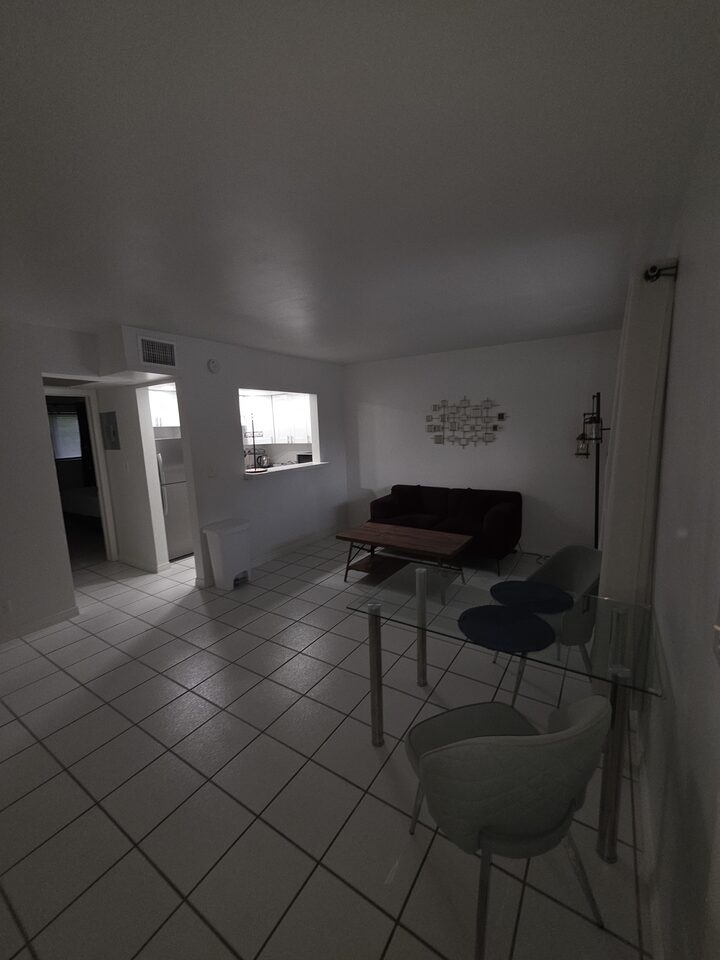 Living and dining area with a burgundy sofa, a glass dining table and the kitchen pass-through, on white tile floors