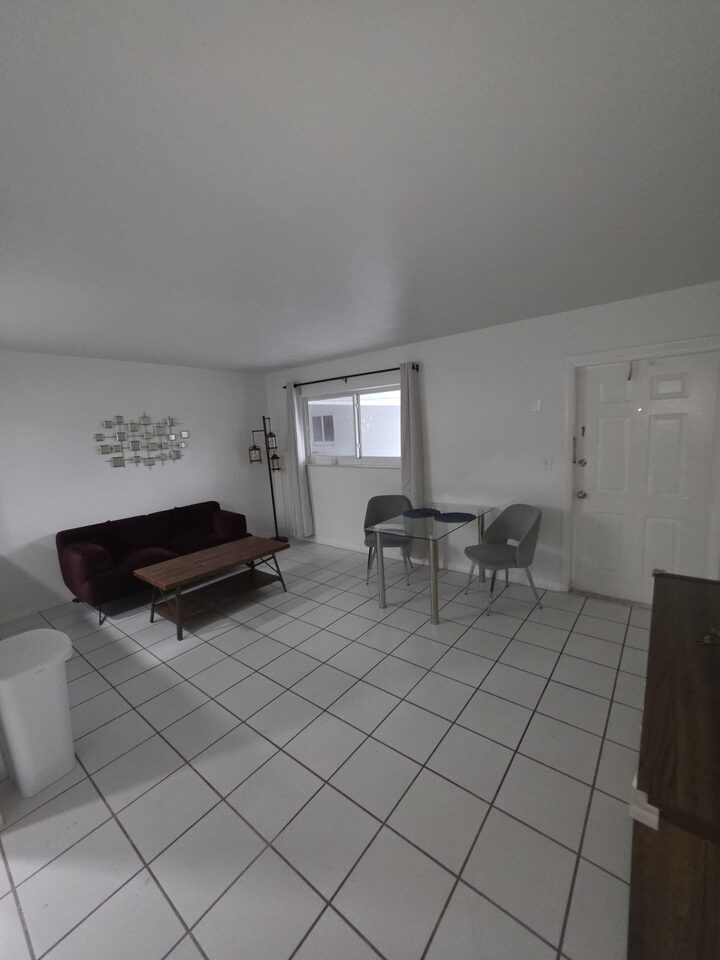 Living room with a burgundy sofa, a wooden coffee table, a glass dining table by the curtained window and the front door beyond