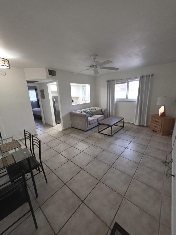 Living room with a grey sofa, a metal-framed coffee table and a ceiling fan, a glass dining table in the foreground, the kitchen pass-through beyond and the bedroom through an open doorway, on large tile floors