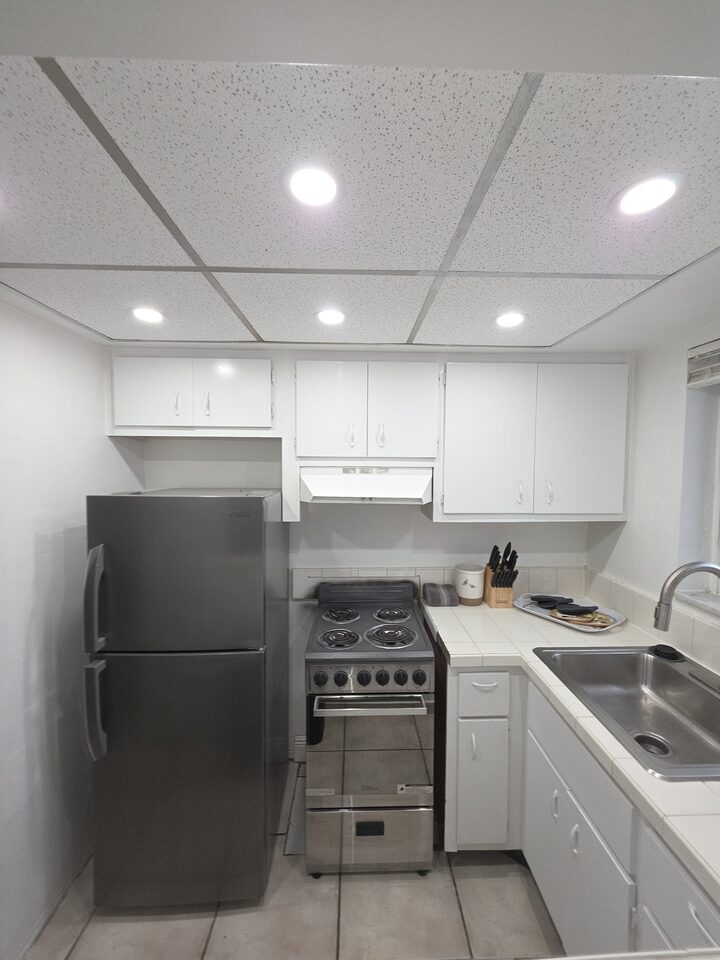 Kitchen with white cabinets, a stainless steel refrigerator, an electric range under an extractor hood, a knife block on a tiled countertop and recessed lights in a panelled ceiling