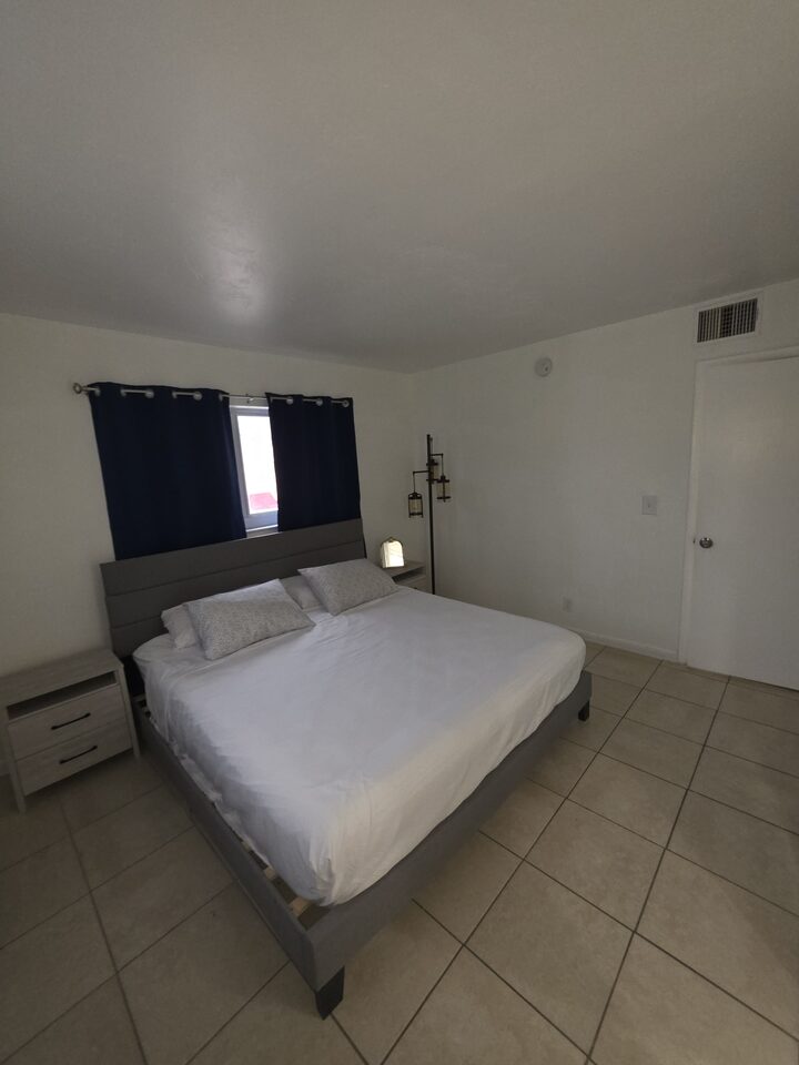 Bedroom seen from the other corner, with the bed, a nightstand, a ceiling air vent and the door to the hallway