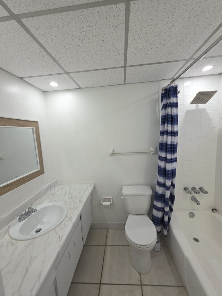 Bathroom with a marble-look vanity and framed mirror, a toilet, and a tub with a rain shower head behind a blue and white curtain