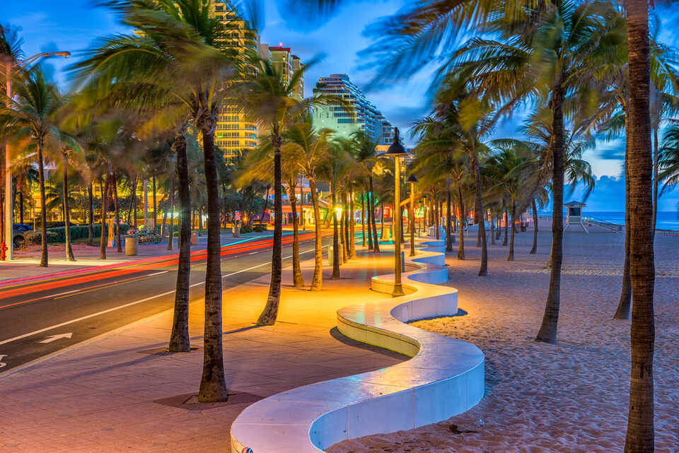 Fort Lauderdale beach at dusk, with palms along the seafront road and the curved white sea wall lit against the sand